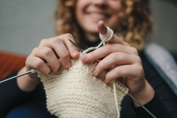 young beautiful happy curly haired woman holding knitting needles knitting clothes for loved ones. f