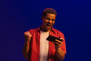 Happy african man gesturing as winner while playing online game on mobile phone isolated over blue neon studio wall