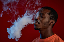 Young Black Man In Orange T-shirt Smoking E-cigarette In Front Of Camera Against Red Background And Exhaling Cloud Of White Smoke