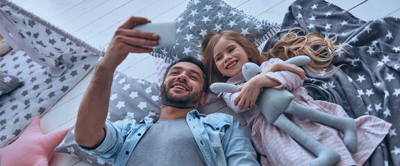 Wall Mural - Top view of happy father and little daughter making selfie while lying on the floor at home together