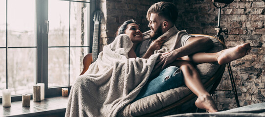 Sticker - Happy young couple embracing while sitting in comfortable armchair at home together