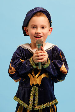 Happy preschool age boy in costume of medieval pageboy, little prince singing at microphone over light blue background. Concept of children emotions, eras comparison, fashion, music