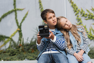 Wall Mural - girl in denim vest and jeans leaning on shoulder of preteen boy in stylish clothes holding vintage camera.