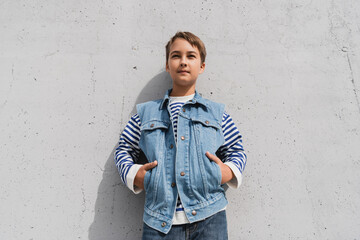 Poster - stylish preteen boy in denim vest and striped long sleeve shirt posing with hands in pockets near mall with grey wall.