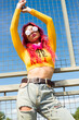 A lady with pink hair, headphones around her neck, in a yellow top, ripped jeans and sunglasses is standing on the street with her hands up. Vintage retro clothing style of the 2000s in modern fashion