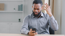 Stressed Mad Angry African American Ethnic Bearded Man With Mobile Phone Reading Bad News. Sad Worker Businessman With Smartphone Failure Online App Error Broken Cellphone Service Problem In Office