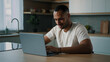 © Yuliia - African millennial man multiracial guy American male user freelancer sitting at home kitchen desk using laptop working distant with computer app service online betting game chatting buying in Internet