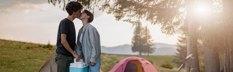 male tourist kissing girlfriend and holding portative fridge at camping site. rural rest.