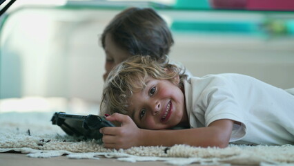 Wall Mural - Portrait of joyful child holding tablet device lying on floor turning head to camera smiling. Kids close up face posing