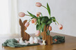 Happy Easter! Beautiful tulips, eggs and bunny decoration on wooden table. Modern farmhouse easter decor. Stylish handmade egg holder, natural eggs, pink tulips bouquet and rustic bunnies