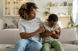 © DimaBerlin - Caring African American mother calms offended child boy while sitting on couch after quarrel at home. Attentive Mom hugs support upset schoolboy with pout lips and crossed hands due to school failures