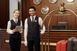 © pressmaster - Happy young elegant receptionist and bellboy standing in lounge of luxurious hotel and looking at camera while waiting for new guests