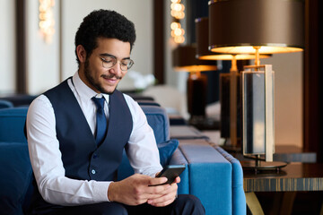 Happy young broker in elegant suit texting in mobile phone or communicating in video chat while sitting in hotel lounge