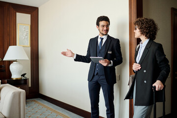 Poster - Young confident head manager of luxurious hotel showing room to female guest in smart casualwear with suitcase listening to him