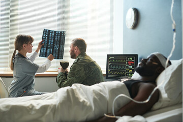 Wall Mural - Nurse in medical scrubs explaining x-ray results to soldier in military uniform while sitting in hospital ward with injured patient