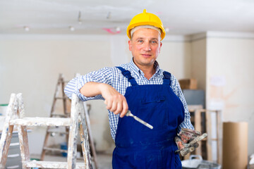 Wall Mural - Portrait of positive man builder holding work tools, standing at stepladder in apartment.