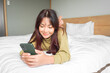 © Reezky - A smiling young Asian woman is laughing while holding her phone and lying on the bed