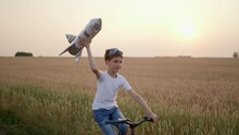 Little Happy Boy Child Teenager Aviator Cosmonaut Riding Bike With Toy Rocket In Sunset Field Summer Nature, Kid Big Dream Flying, Astronaut, Space. Concept Of Success Winner Travel Freedom Childhood 