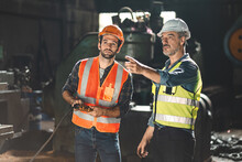 Senior Male Engineer Training And Explaining Work To New Employee Wearing Vest And Safety Jacket With Hardhat Helmet While Pointing Towards Machine In Factory And Giving Instructions