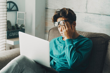 Tired young boy student studying at home sitting on the sofa using laptop and touching his eyes. Stressed education leisure indoor activity. Online class school. Headache teenager study activity