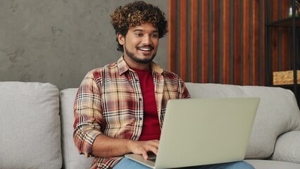Smart IT user young Indian man wears casual clothes hold use work typing on laptop pc computer sits on grey sofa couch stay at home hotel flat rest relax spend free spare time in living room indoor