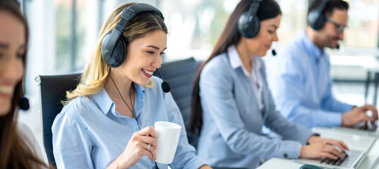 Wall Mural - Wide view of beautiful blonde girl with coffee cup working on computer accompanied with group of call center operators.