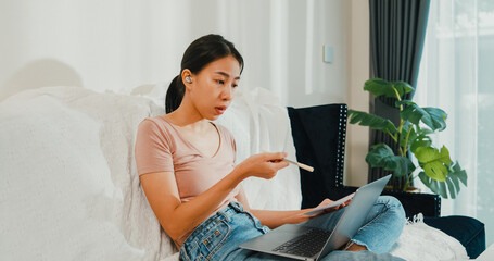 Wall Mural - Asia businesswoman using laptop talk to colleagues about plan in video call while working from house at living room. Remotely at workplace, social distancing, quarantine for corona virus prevention.