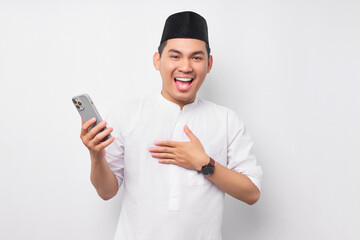 Wall Mural - Smiling happy handsome young Asian Muslim man in Arabic clothes holding mobile phone and receiving good news isolated on white background. People religious Islamic lifestyle concept