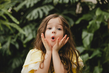 Little girl in a yellow dress in a botanical garden. a child stands near the leaves of Monstera.