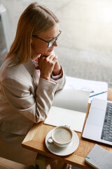 Wall Mural - Woman freelancer working on laptop and drinking coffee while sitting in cafe near window