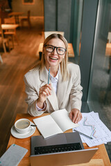Wall Mural - Smiling businesswoman drinking coffee and making notes while working on laptop in cafe