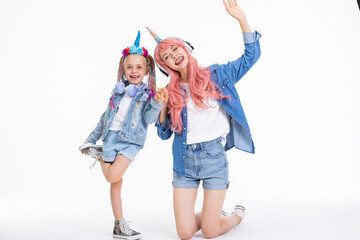 Happy adorable mother with pink wig and unicorn headband hugging preschooler daughter with braids standing on white background in studio isolaed in denim clothes having fun posing fo camera.