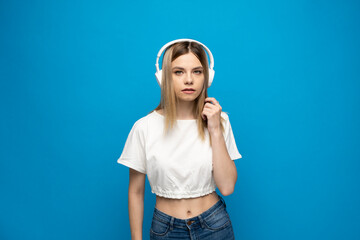 Cheerful young blonde woman girl in white clothes listening music with headphones, mobile phone isolated on blue background studio. People lifestyle concept.