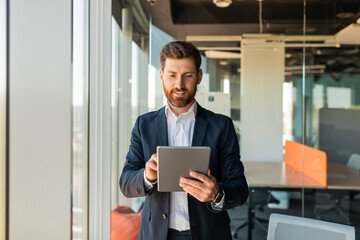 Poster - Happy male entrepreneur using digital tablet and smiling, standing near window at modern coworking office, copy space