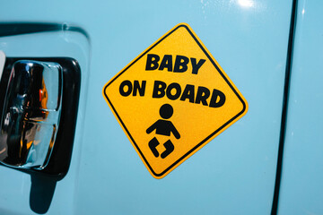 Close up on a yellow sticker in the shape of a warning sign with the inscription 'Baby on Board' stuck to the back of a turquoise colored car