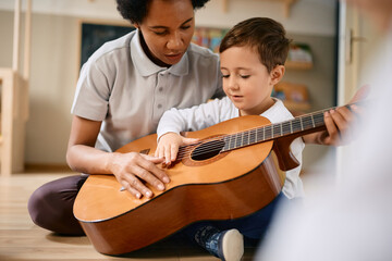 Wall Mural - Small boy learns to play acoustic guitar with help of his African American teacher at kindergarten.