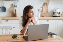 Pensive Hispanic Young Adult Woman Sits At Table With Laptop At Kitchen Exhausted Of Hard Work. Sad Italian Girl Feels Fatigue Looks Aside Thinks About Financial Troubles. Overloaded Female, Failure.