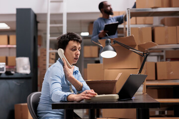 Wall Mural - Caucasian manager talking at landline phone with remote supervisor discussing transportation logistics problem while looking at cargo stock on laptop. Employee working at order delivery in warehouse