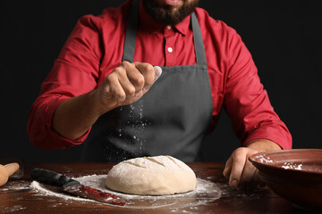 Canvas Print - Male baker sprinkling dough with flour at table on dark background, closeup