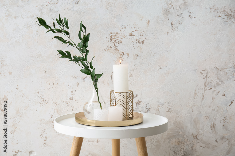 Burning candles and vase with ruscus branch on table near grey grunge wall
