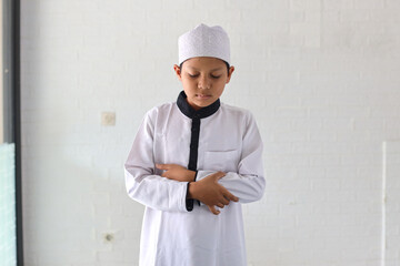 Wall Mural - Asian muslim kid wearing muslim rob and skullcap stands with hand in chest, praying salah on prayer mat.