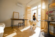 © rh2010 - Young woman stands with her dog by the window blinds and looks away in cozy and sunny living room of modern apartment in beige tones. Quarantine, loneliness and life at home concept