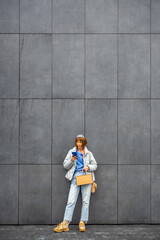  Young stylish woman uses mobile phone while standing on background of gray wall outdoors. Wide view with copy space