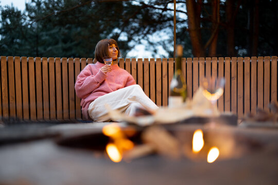 young woman sits with a wine on round bench while resting at beautiful bbq area illuminated with gar