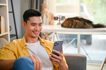 Handsome Asian man using his smartphone while relaxing on sofa in his living room.