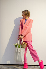 Lifestyle portrait of stylish woman in pink suit with eco handbag full of flowers on white background. Canvas bag with blank space. Concept of style and spring time