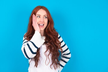 Wall Mural - young caucasian woman wearing overalls over blue background excited looking to the side hand on face. Advertisement and amazement concept.
