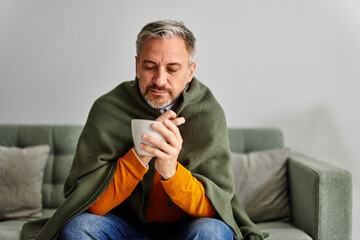 A sick bearded man wrapped in a blanket having a cup of warm tea at home.