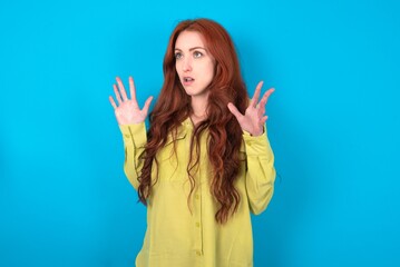 Wall Mural - young woman wearing green sweater over blue background shouts loud, keeps eyes opened and hands tense.