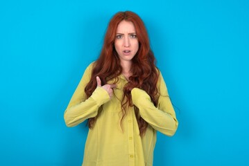 Wall Mural - Embarrassed young woman wearing green sweater over blue background indicates at herself with puzzled expression, being shocked to be chosen to participate in competition, hesitates about something